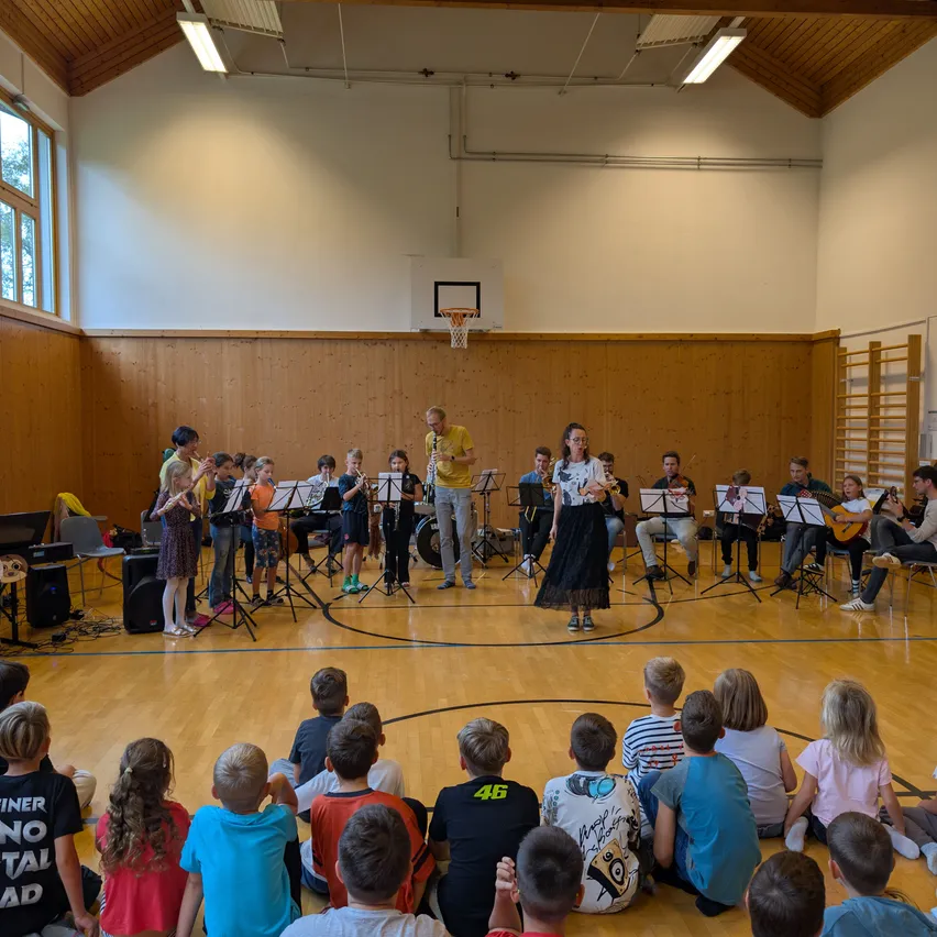 Eine Gruppe von Kindern und Erwachsenen versammelt sich in einer Turnhalle und spielt verschiedene Musikinstrumente. Die Kinder sitzen auf dem Boden, während die Erwachsenen auf dem Basketballplatz stehen. Eine Frau in einem schwarzen Kleid spielt Geige.