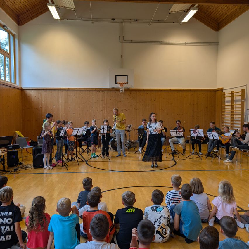 Eine Gruppe von Kindern und Erwachsenen versammelt sich in einer Turnhalle und spielt verschiedene Musikinstrumente. Die Kinder sitzen auf dem Boden, während die Erwachsenen auf dem Basketballplatz stehen. Eine Frau in einem schwarzen Kleid spielt Geige.