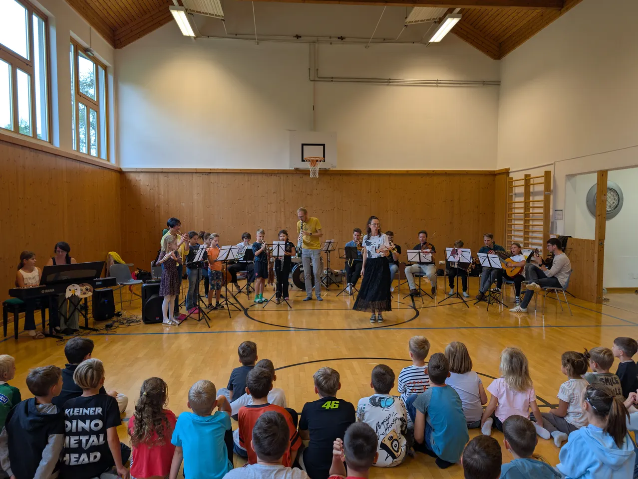 Eine Gruppe von Kindern und Erwachsenen versammelt sich in einer Turnhalle und spielt verschiedene Musikinstrumente. Die Kinder sitzen auf dem Boden, während die Erwachsenen auf dem Basketballplatz stehen. Eine Frau in einem schwarzen Kleid spielt Geige.