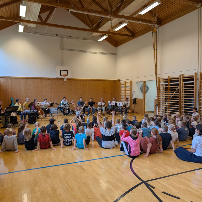 Eine Gruppe von Musikern tritt in einer Turnhalle auf, umgeben von einem großen Publikum aus Kindern, die auf dem Boden sitzen. Die Musiker spielen verschiedene Instrumente, darunter Celli, Schlagzeug und Klavier. Die Turnhalle hat Holzwände und einen Basketballkorb.