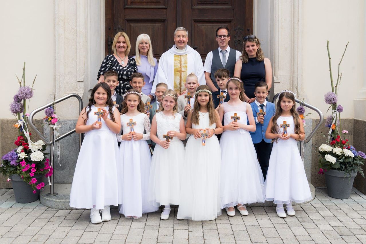 Eine Gruppe von Kindern und Erwachsenen in weißen Kleidern steht vor einer Kirche und hält Kreuze. Ein Priester steht in der Mitte, und zwei Frauen stehen hinter ihm.