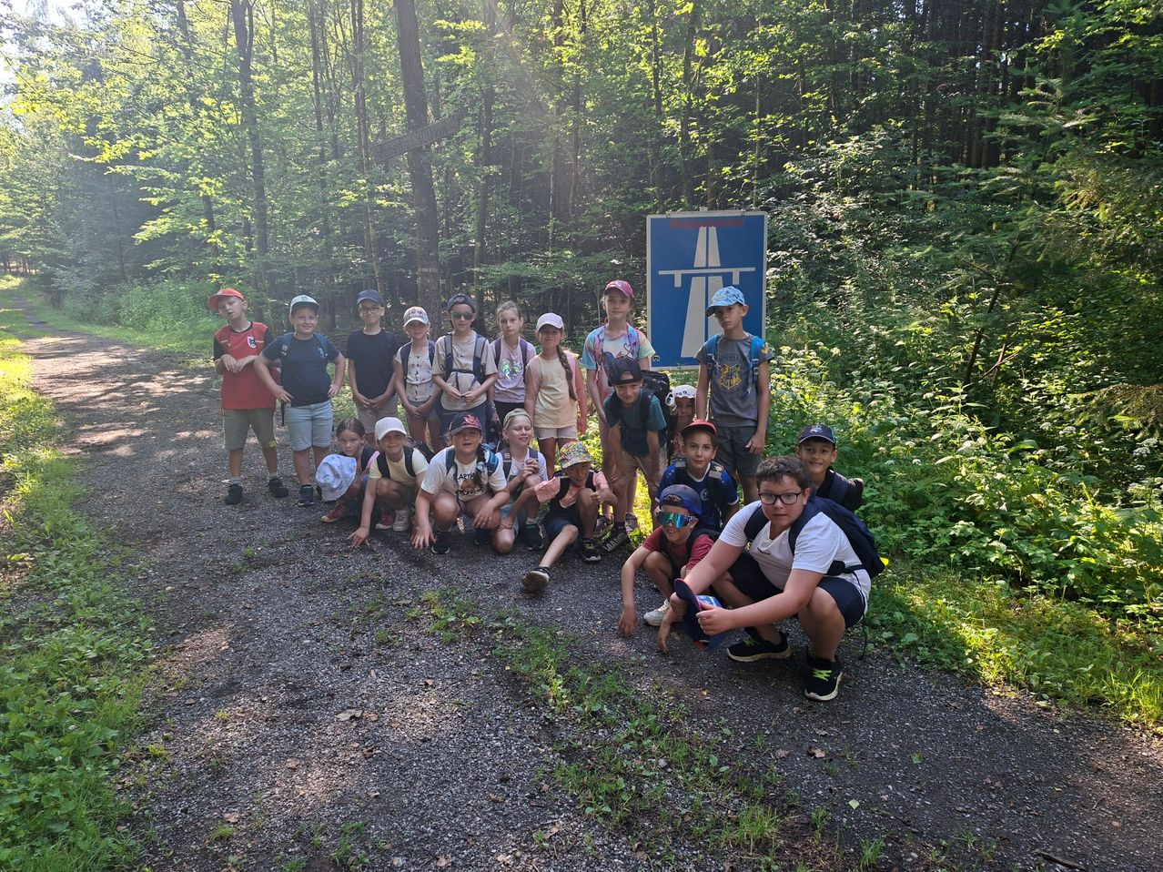 Eine Gruppe junger Leute mit Hüten und Rucksäcken posiert für ein Foto auf einem Schotterweg mit einem blauen Schild im Hintergrund.