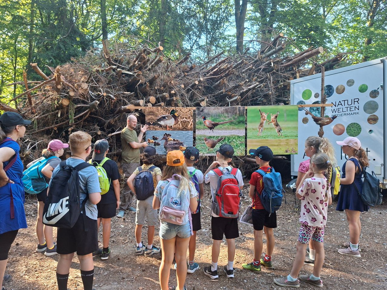 Eine Gruppe von Kindern und einem Erwachsenen sind um einen Mann versammelt, der ihnen Bilder von Vögeln auf einem Bildschirm zeigt. Die Kinder tragen Rucksäcke und scheinen aufmerksam zuzuhören. Im Hintergrund befindet sich ein Haufen von Baumstämmen und eine weiße Struktur mit einem grünen Kreisdesign.