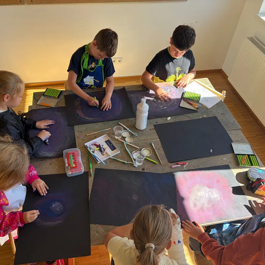 Eine Gruppe von Kindern sitzt um einen Tisch herum und zeichnet auf schwarzes Papier mit verschiedenen Malutensilien.