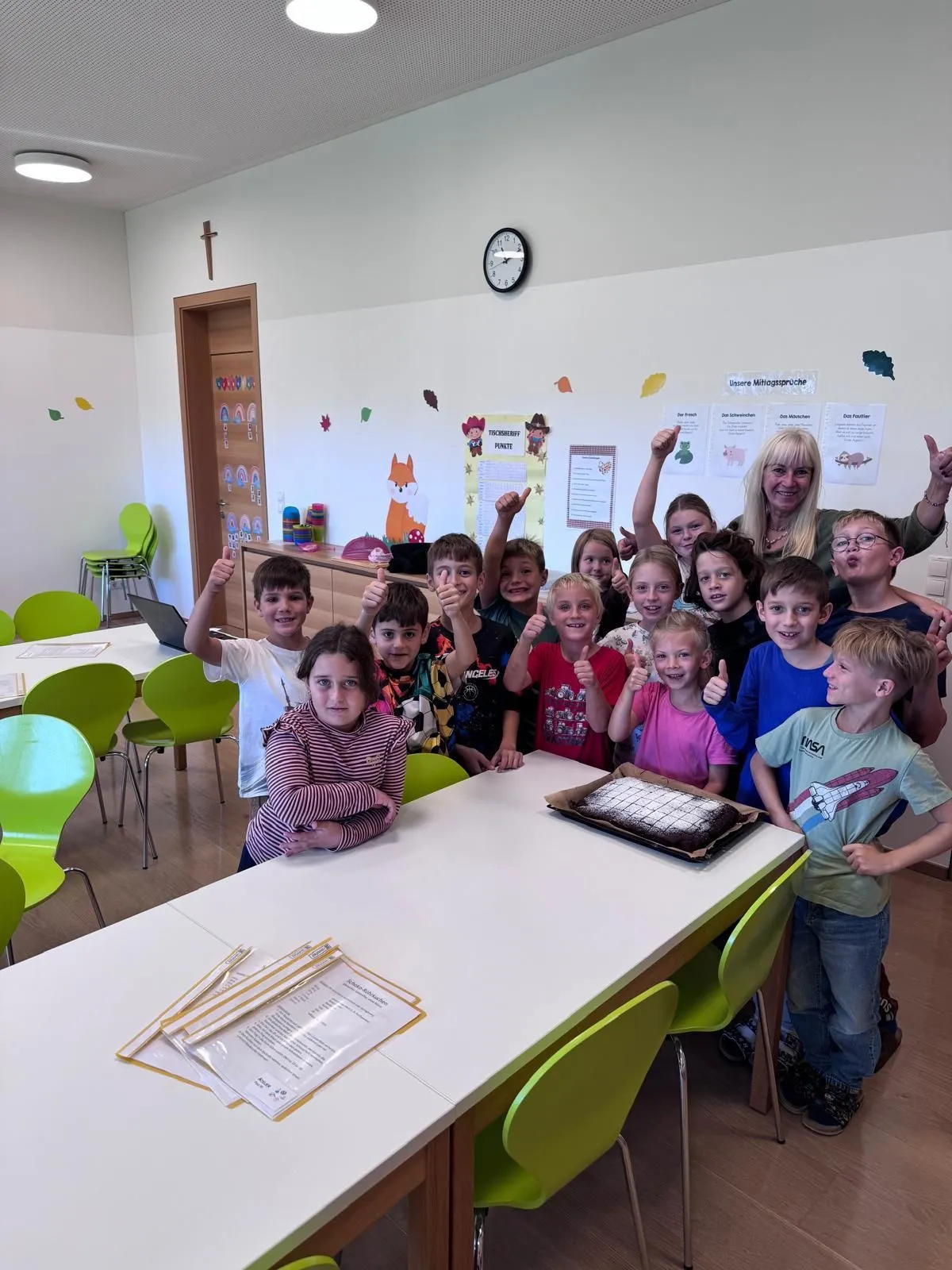 Eine Gruppe von Kindern und einer erwachsenen Frau sind in einem Klassenzimmer. Sie posieren für ein Foto mit einem Kuchen auf dem Tisch.