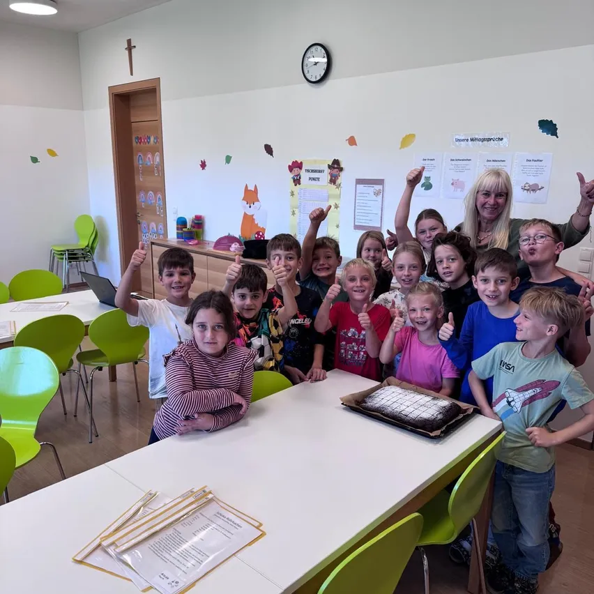 Eine Gruppe von Kindern und einer erwachsenen Frau sind in einem Klassenzimmer. Sie posieren für ein Foto mit einem Kuchen auf dem Tisch.