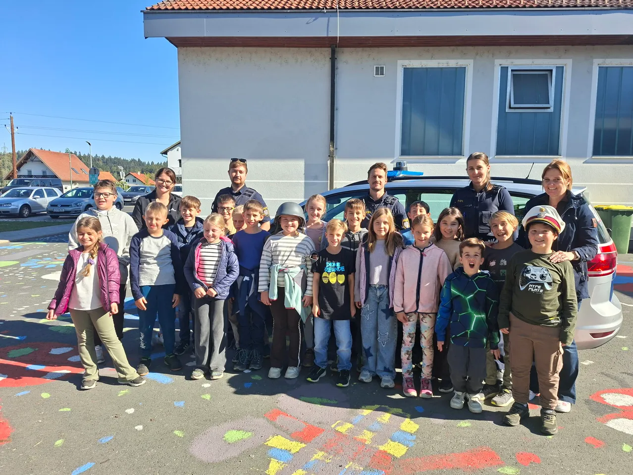 Eine Gruppe von Kindern und Erwachsenen, darunter zwei Polizeibeamte, steht vor einem Gebäude mit einer farbenfrohen Kreidezeichnung auf dem Boden.