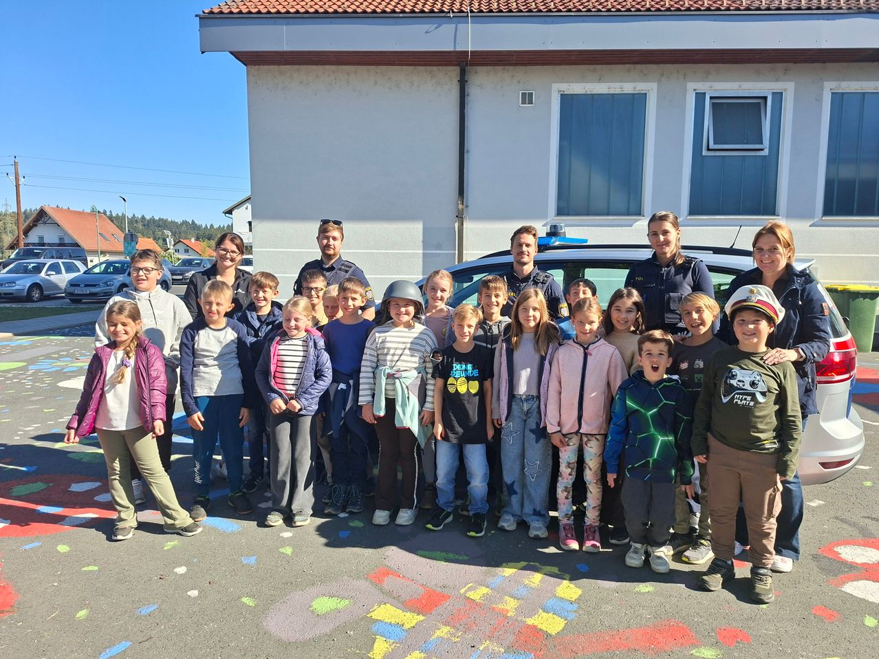 Eine Gruppe von Kindern und Erwachsenen, darunter zwei Polizeibeamte, steht vor einem Gebäude mit einer farbenfrohen Kreidezeichnung auf dem Boden.