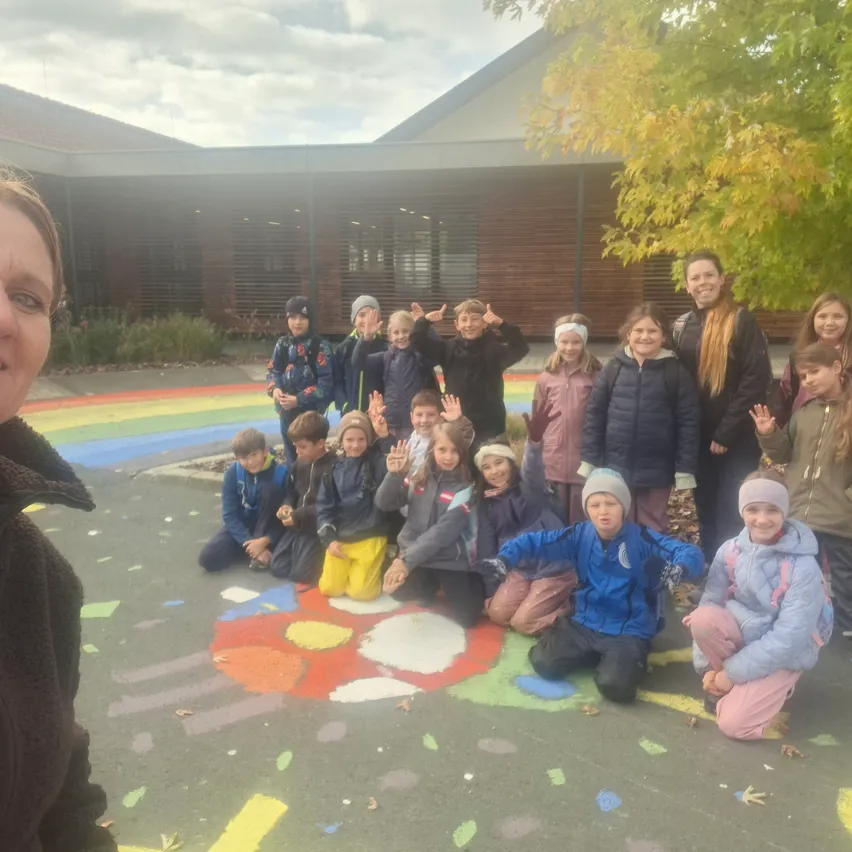 Eine Gruppe von Kindern und einer erwachsenen Frau posiert für ein Foto vor einem Gebäude. Die Kinder sitzen auf dem Boden und lächeln. Die erwachsene Frau steht auf der linken Seite.