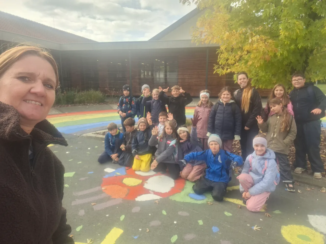 Eine Gruppe von Kindern und einer erwachsenen Frau posiert für ein Foto vor einem Gebäude. Die Kinder sitzen auf dem Boden und lächeln. Die erwachsene Frau steht auf der linken Seite.