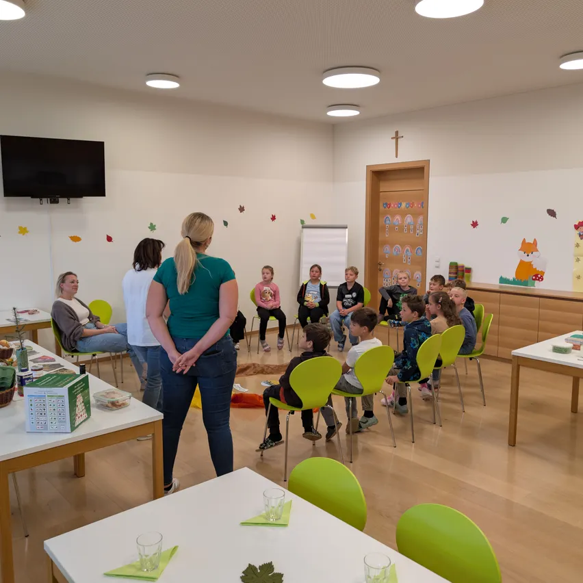 Eine Gruppe von Kindern sitzt im Kreis auf grünen Stühlen in einem Klassenzimmer. Eine Frau steht vor ihnen, und ein Tisch mit Essen steht links.