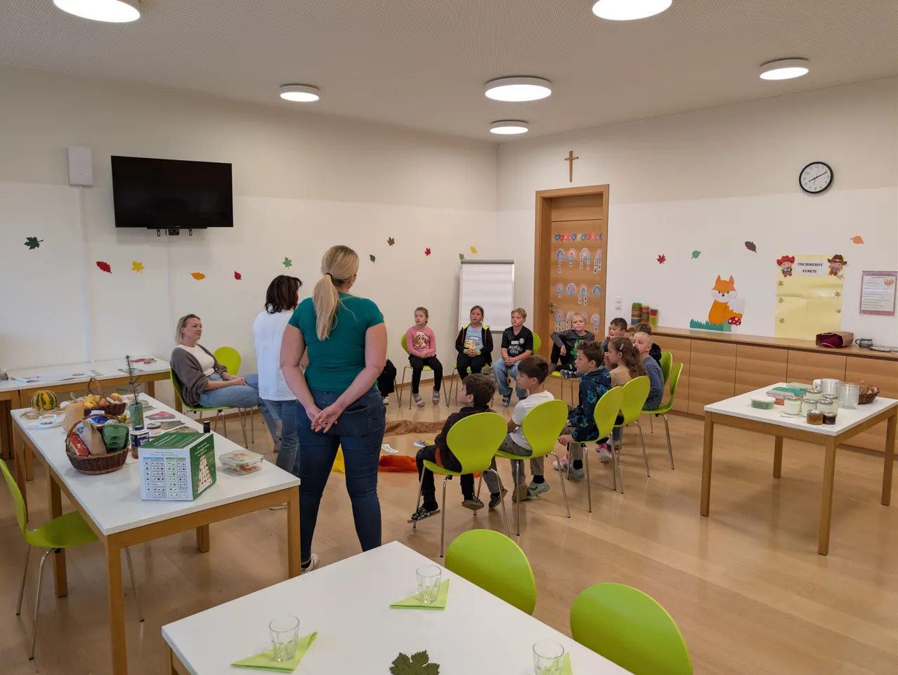 Eine Gruppe von Kindern sitzt im Kreis auf grünen Stühlen in einem Klassenzimmer. Eine Frau steht vor ihnen, und ein Tisch mit Essen steht links.