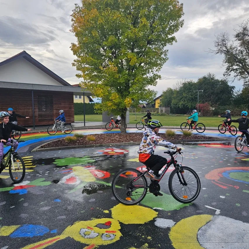 Eine Gruppe von Kindern fährt auf einer bunt bemalten Straße mit Fahrrädern, mit einem Gebäude und Bäumen im Hintergrund.