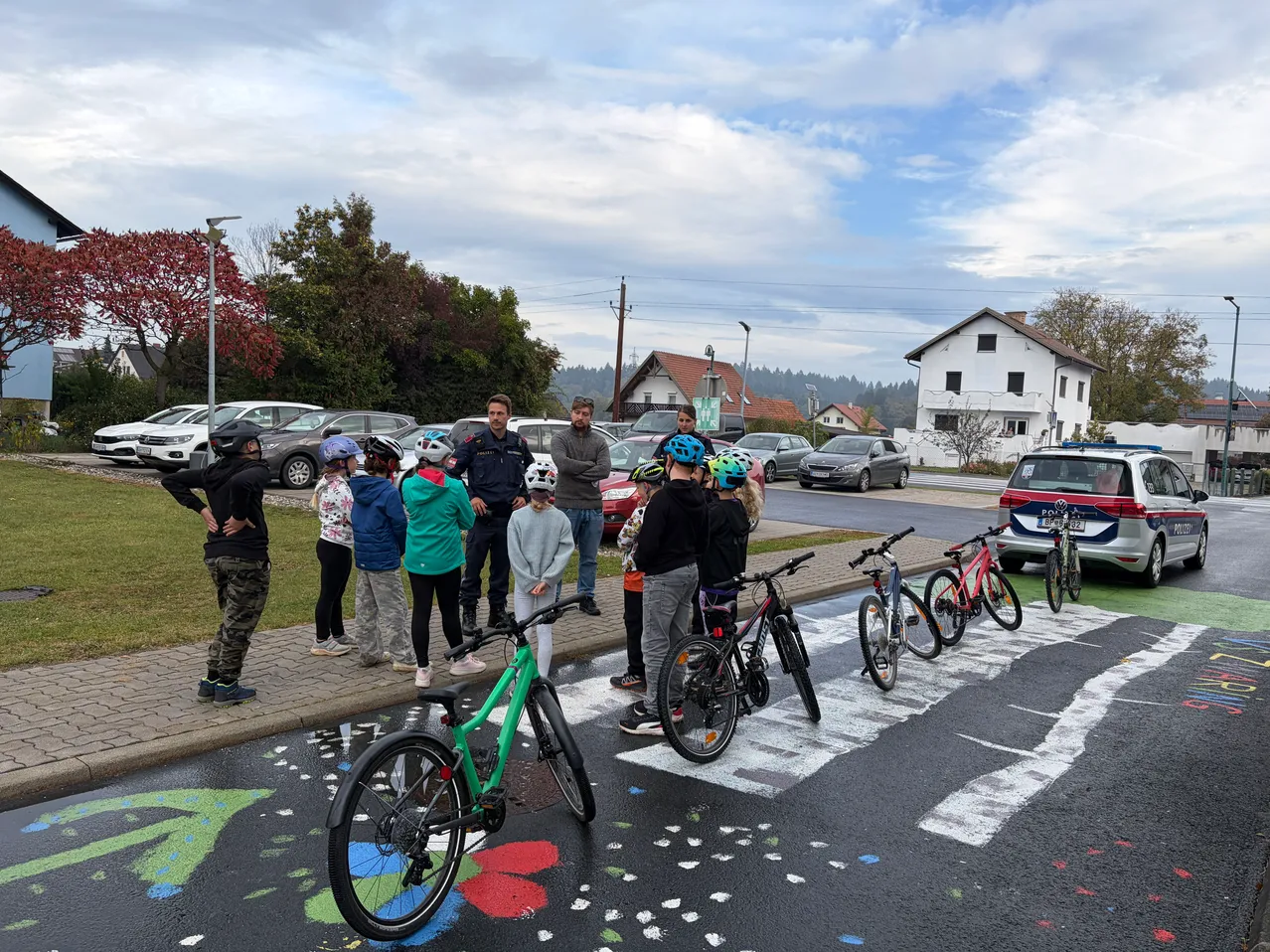 Eine Gruppe von Menschen, einige mit Helmen, versammelt sich auf einer Straße mit bemalten Fahrrädern und einem Polizeiwagen in der Nähe.