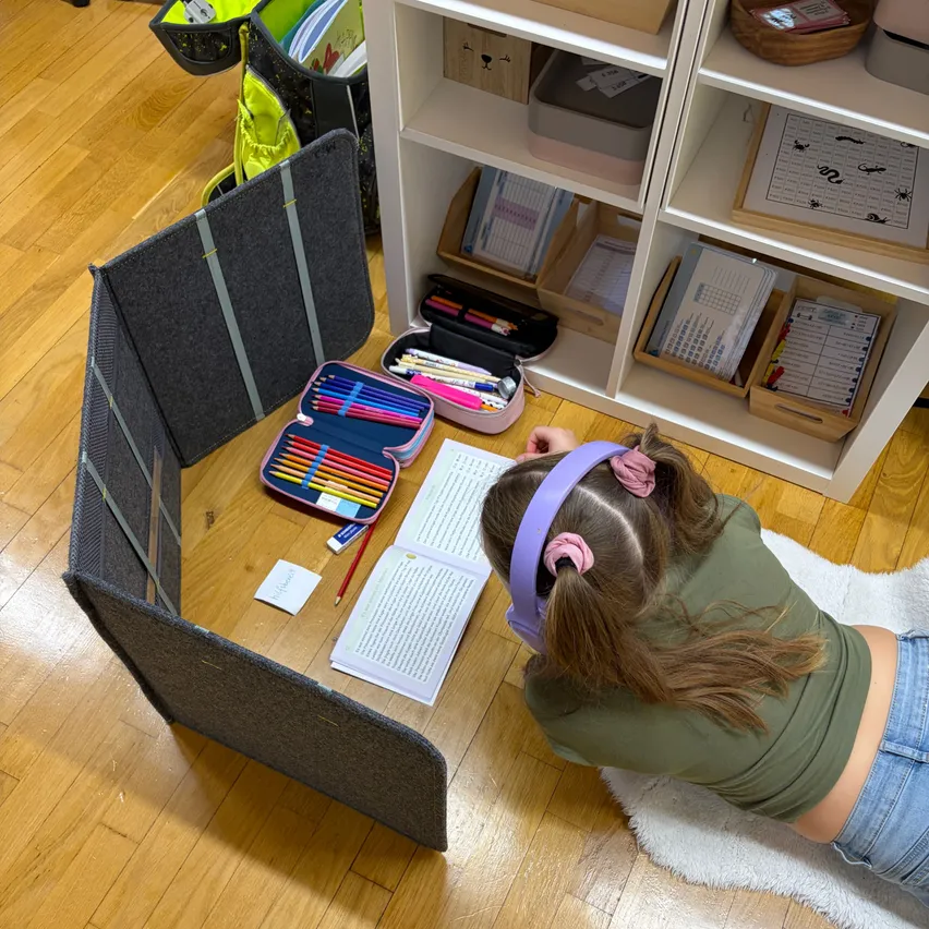 Ein junges Mädchen mit Kopfhörern liegt auf dem Boden und studiert ein Buch. Sie hat einen Stiftetui mit farbigen Stiften und ein Notizbuch auf dem Tisch vor sich.
