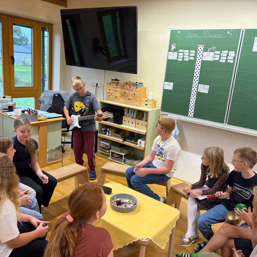 Eine Gruppe von Kindern sitzt um einen Tisch in einem Klassenzimmer. Ein Junge spielt Gitarre, während ein anderer Junge auf einem Stuhl sitzt und zuhört.