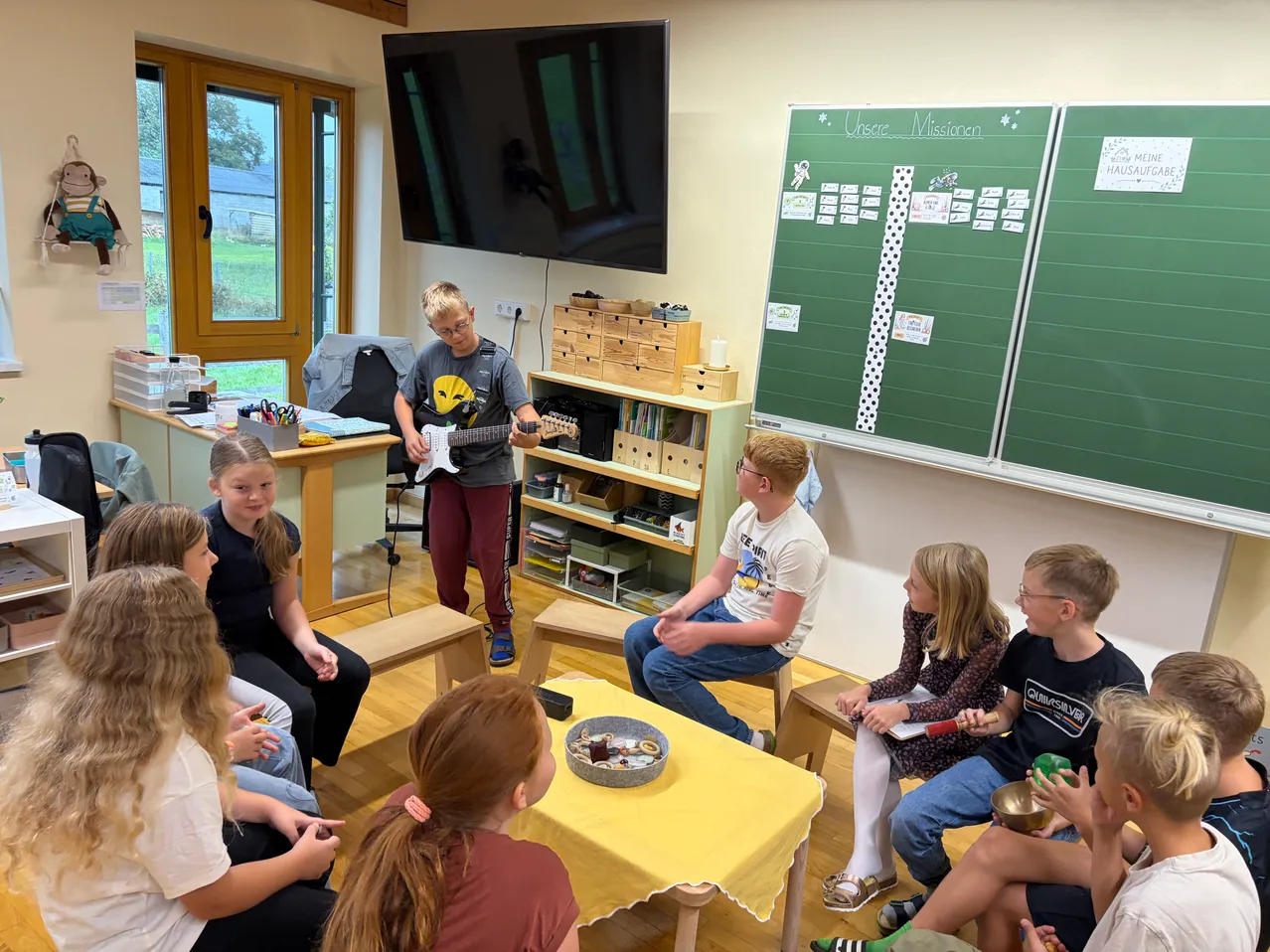 Eine Gruppe von Kindern sitzt um einen Tisch in einem Klassenzimmer. Ein Junge spielt Gitarre, während ein anderer Junge auf einem Stuhl sitzt und zuhört.