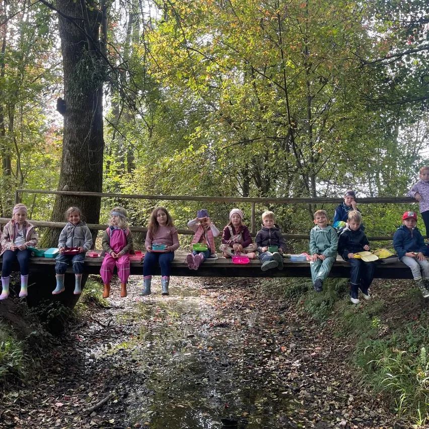 Eine Gruppe von Kindern sitzt auf einer Holzbrücke im Wald und hält Teller mit Essen.