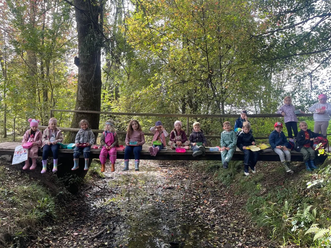 Eine Gruppe von Kindern sitzt auf einer Holzbrücke im Wald und hält Teller mit Essen.