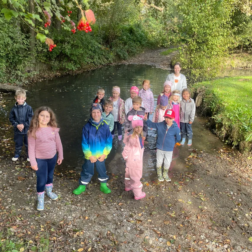 Eine Gruppe von Kindern und einer erwachsenen Frau steht in einem Bach mit Wasser bis zu den Knien. Sie tragen alle Winterkleidung und Stiefel.