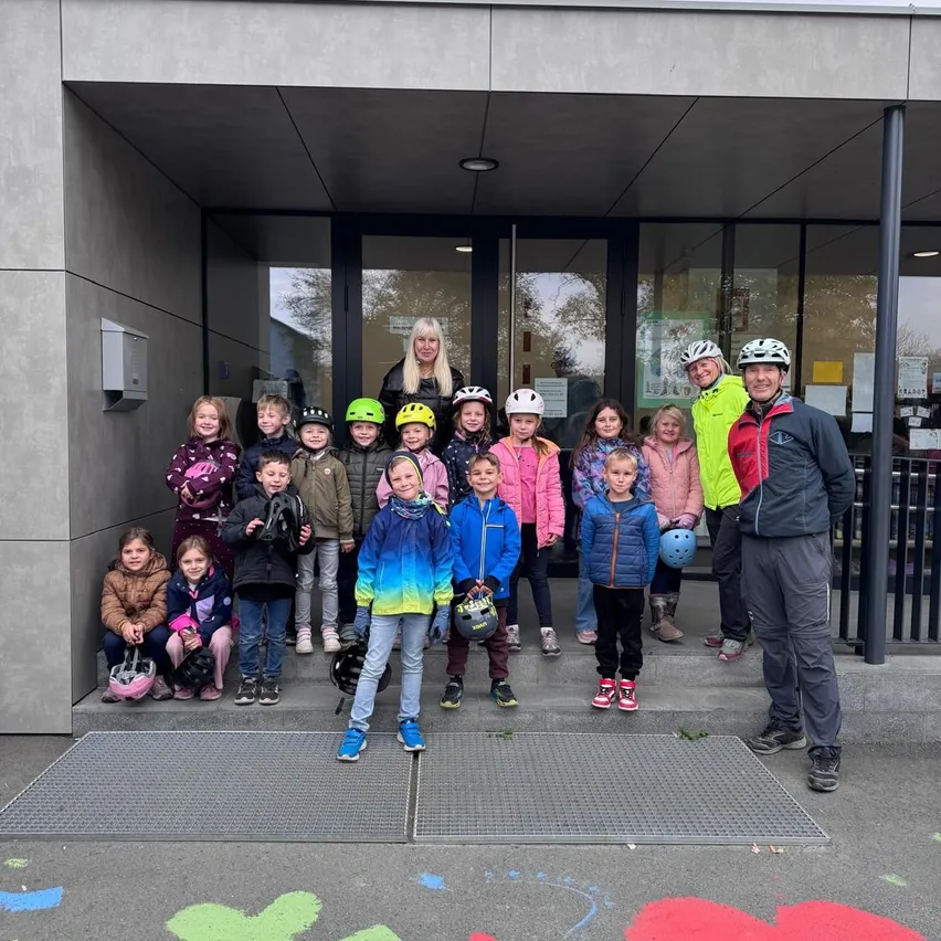 Eine Gruppe von Kindern und Erwachsenen steht vor einem Gebäude. Die Kinder tragen Helme und halten sie in ihren Händen.