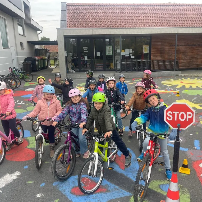 Eine Gruppe von Kindern steht mit ihren Fahrrädern auf einem Parkplatz. Sie tragen alle Helme und Jacken. Der Parkplatz hat bunte Zeichnungen auf dem Boden.