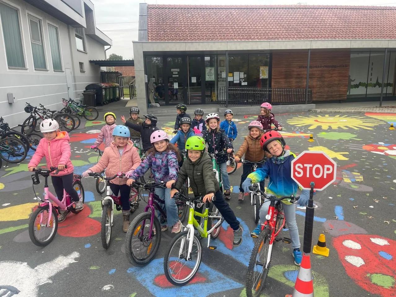Eine Gruppe von Kindern steht mit ihren Fahrrädern auf einem Parkplatz. Sie tragen alle Helme und Jacken. Der Parkplatz hat bunte Zeichnungen auf dem Boden.