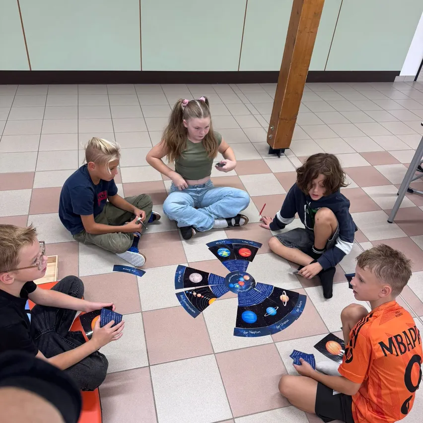 Fünf Kinder sitzen auf dem Boden in einem Klassenzimmer und spielen ein Spiel mit einer Sonnensystemtafel.