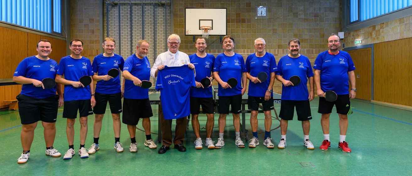 Eine Gruppe von Männern in blauen Shirts und schwarzen Shorts posiert für ein Foto in einer Turnhalle, Paddles in der Hand. Ein Mann in einem weißen Hemd hält ein blaues Shirt mit Text. Hinter ihnen steht ein Basketballkorb, und eine Uhr ist an der Wand montiert.