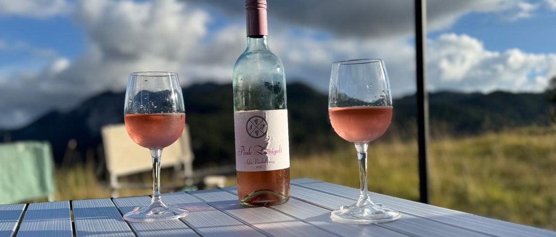Eine Flasche Pink Zweigelt steht zwischen zwei mit rosa Wein gefüllten Gläsern auf einem Tisch mit einer malerischen Kulisse im Hintergrund.