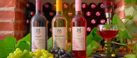 Three bottles of wine and a glass of wine sit on a table with grapes and a brick wall in the background.