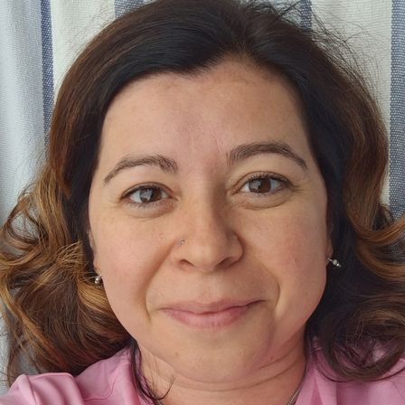 Eine Frau mit braunen Haaren und einem rosa Shirt lächelt für ein Selfie. Sie hat eine Nasenpiercing und Ohrringe. Der Hintergrund ist unscharf.