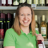 Eine Frau in einem grünen Shirt lächelt und hält eine Flasche Wein. Hinter ihr befinden sich Regale mit verschiedenen Flaschen Wein.