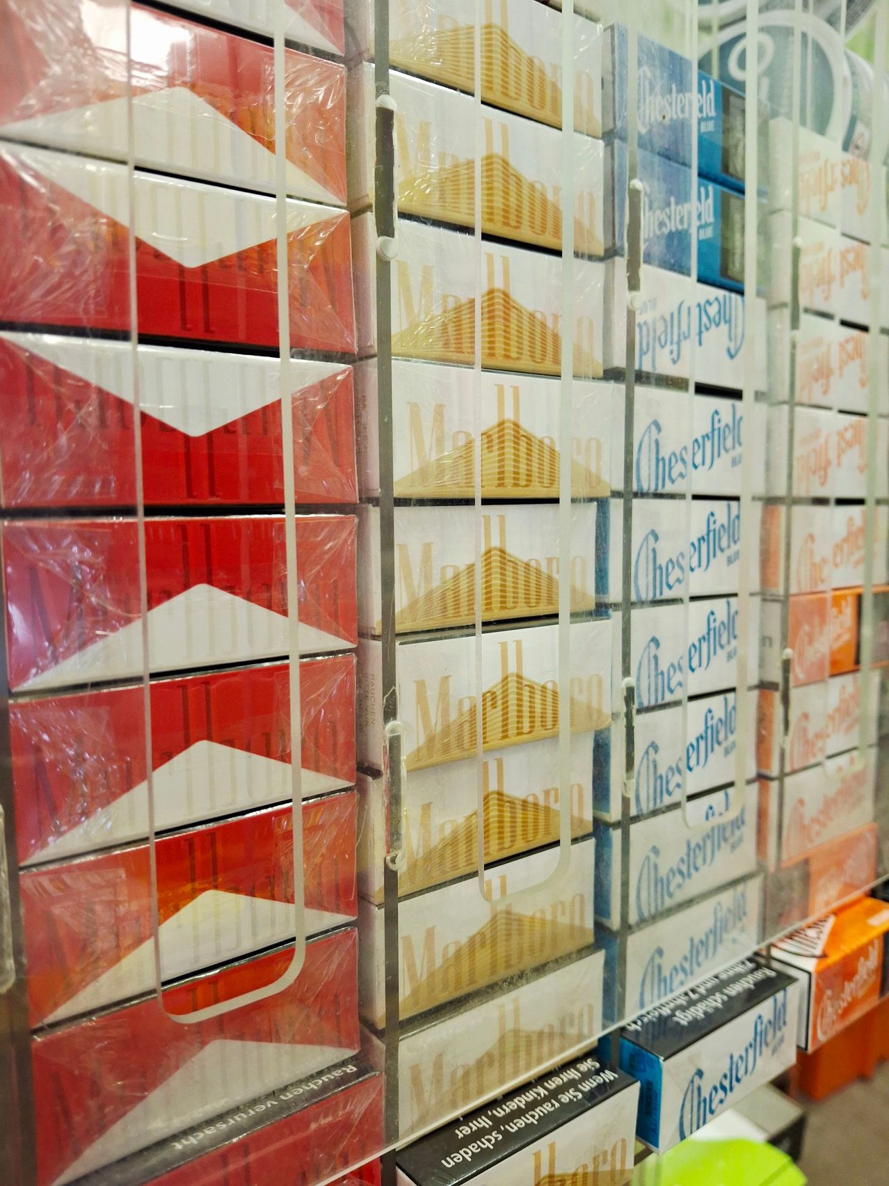 Eine Reihe von Zigarettenpackungen, einschließlich Marlboro und Chesterfield, sind in einem Geschäft gestapelt. Die Marlboro-Packungen sind in Rot, Weiß und Gelb, während die Chesterfield-Packungen in Blau und Weiß sind. Alle sind mit Plastik bedeckt.