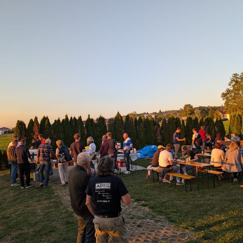 Eine Gruppe von Menschen versammelt sich auf einem Grasfeld mit Tischen und Bänken. Einige sitzen, andere stehen. Bäume und ein Feld sind im Hintergrund.