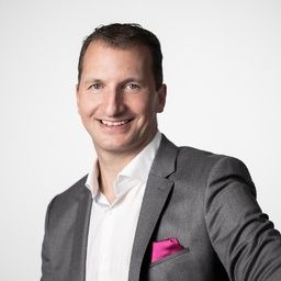 Ein Mann in einem grauen Anzug und einem weißen Hemd mit einem rosa Einstecktuch im Revers lächelt und posiert für ein Foto.