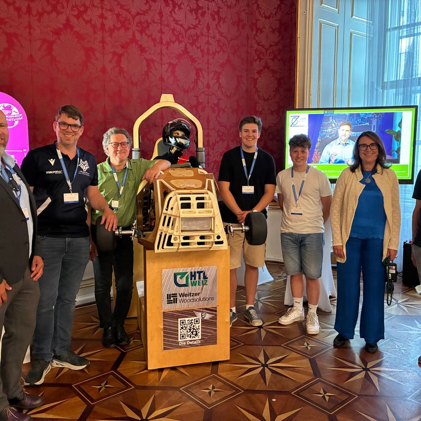 Eine Gruppe von Menschen posiert mit einem hölzernen Automodell und einem Helm in einem Raum mit einem Bildschirm, der einen Mann zeigt.