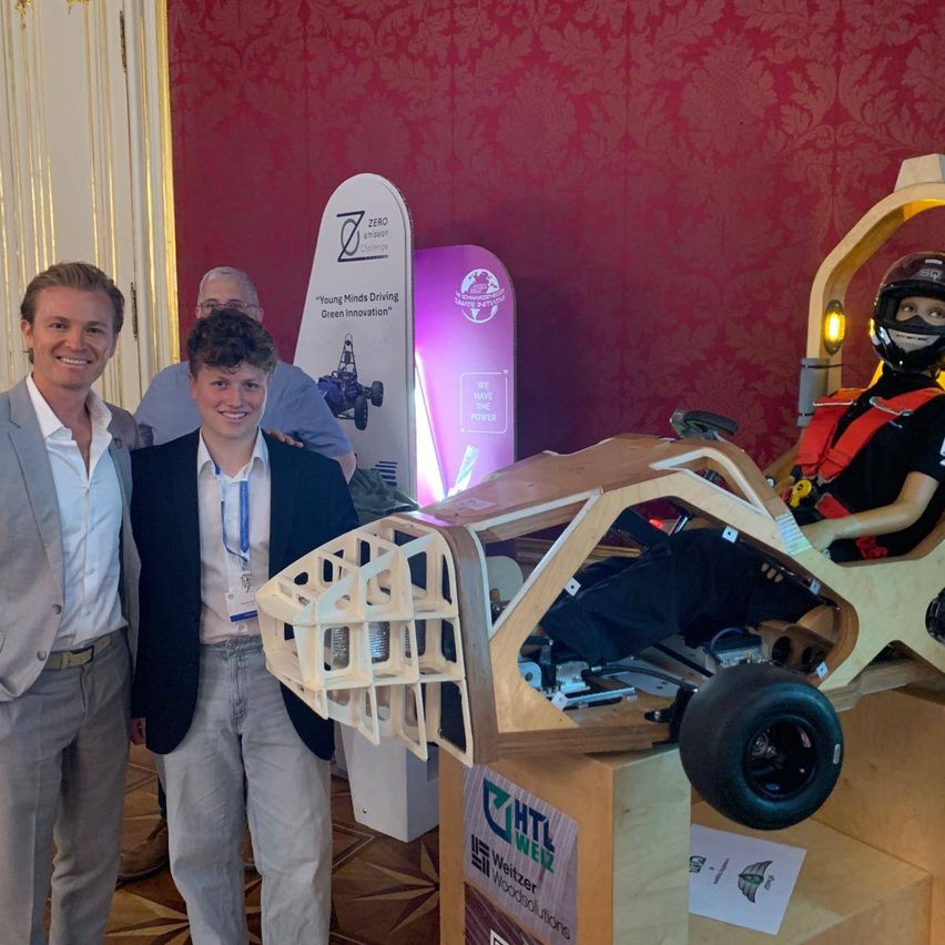 Drei Personen stehen vor einem hölzernen Fahrzeug, wobei eine einen Helm trägt und eine andere ein weißes Schild hält. Dahinter zeigt eine rote Wand ein Schild mit den Worten 'ZERO' und 'Saubere Energie'.