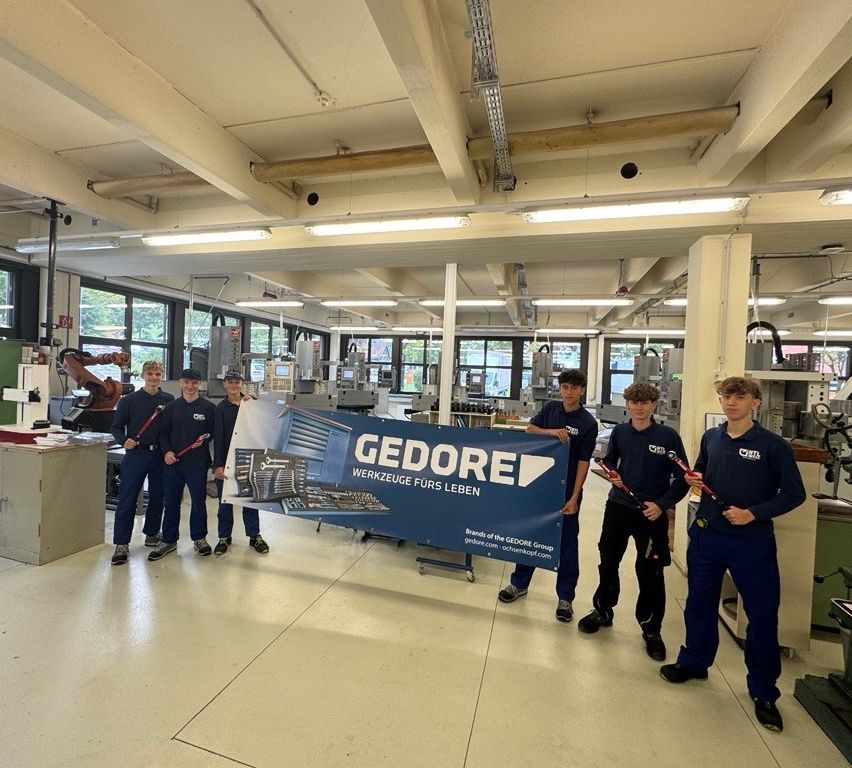 Fünf Personen in blauen Uniformen posieren mit einem großen Banner in einer Fabrik. Das Banner trägt die Aufschrift 'GEDORE' mit zusätzlichem Text auf Deutsch.