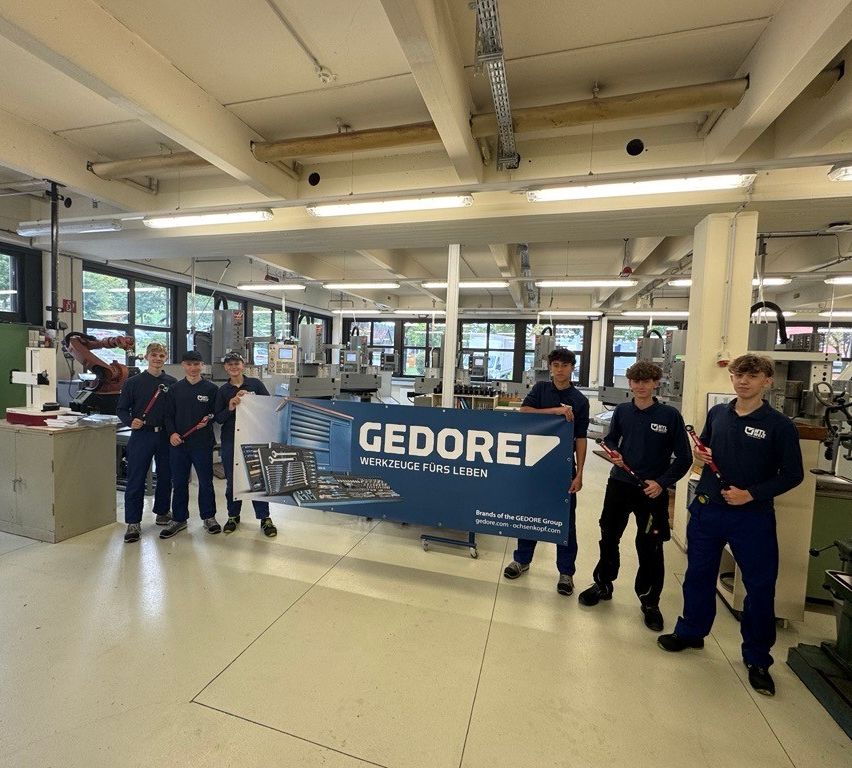Eine Gruppe junger Arbeiter in blauen Uniformen steht in einer Fabrik und hält Werkzeuge und ein Banner. Das Banner trägt den Schriftzug 'GEDORE' und es befinden sich Maschinen im Hintergrund.