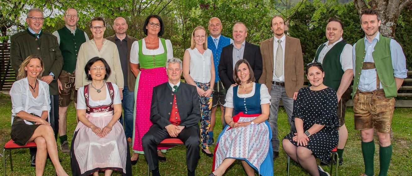 Eine Gruppe von Menschen in traditioneller deutscher Kleidung posiert für ein Foto an einem sonnigen Tag auf einer Rasenfläche mit Bäumen und Bergen im Hintergrund.