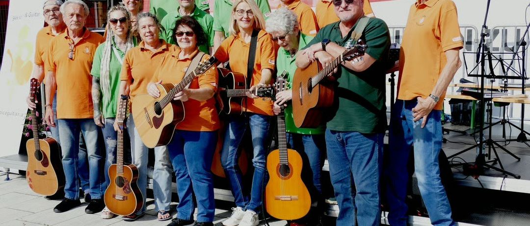 Eine Gruppe von Erwachsenen, sowohl Männer als auch Frauen, posiert für ein Foto. Sie halten Gitarren und tragen orange und grüne Shirts. Sie lächeln und stehen vor einer Bühne.
