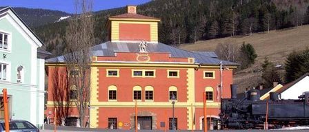 Ein altes, rotes und gelbes Gebäude mit mehreren Fenstern steht neben einem Zug in einem Gebirgsgebiet. Das Gebäude hat einen Turm und eine Statue auf der Spitze. Bäume umgeben das Gebiet.