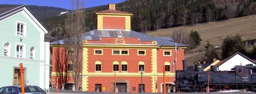 Ein großes rotes und gelbes Gebäude mit vielen Fenstern steht in einer Stadt. Ein Lokomotive ist davor geparkt. Berge sind im Hintergrund.