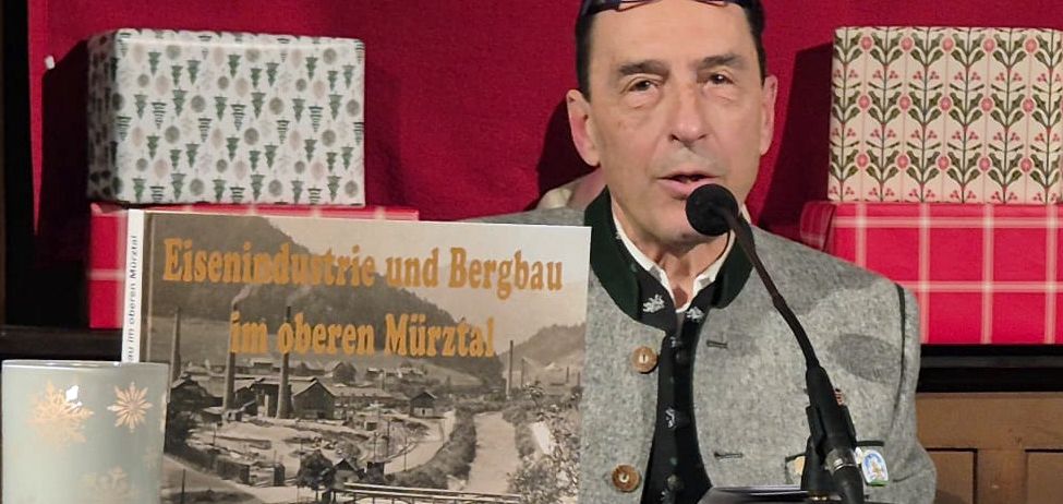 A man speaks into a microphone at an event. He holds a book titled 'Eisenindustrie und Bergbau im oberen Mürztal'. The background features wrapped gifts.