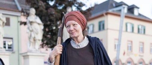 Eine Person in mittelalterlicher Kleidung, mit einem Holzstab und einer Laterne, steht auf einer Stadtstraße.
