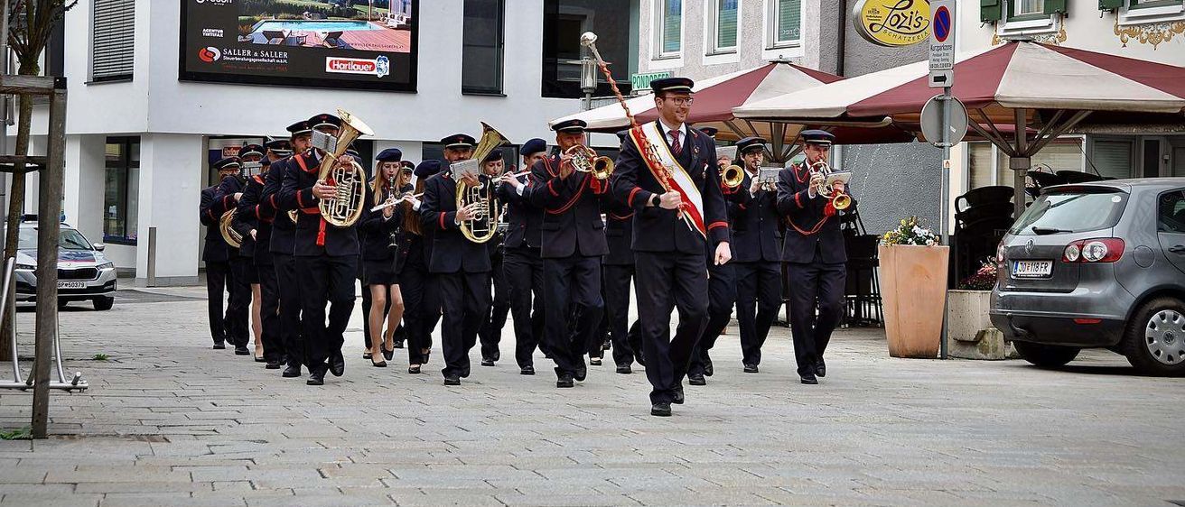 Eine Blaskapelle spielt auf einer Stadtstraße vor einem Gebäude mit einer großen Leinwand.