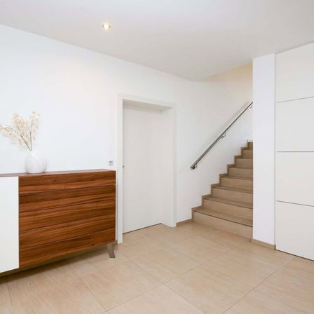 Ein modernes minimalistisches Interieur mit weißen Wänden, einem Holzschrank und einer Treppe, die in den oberen Stock führt. Ein weißer Vase mit getrockneten Blumen steht auf dem Schrank.