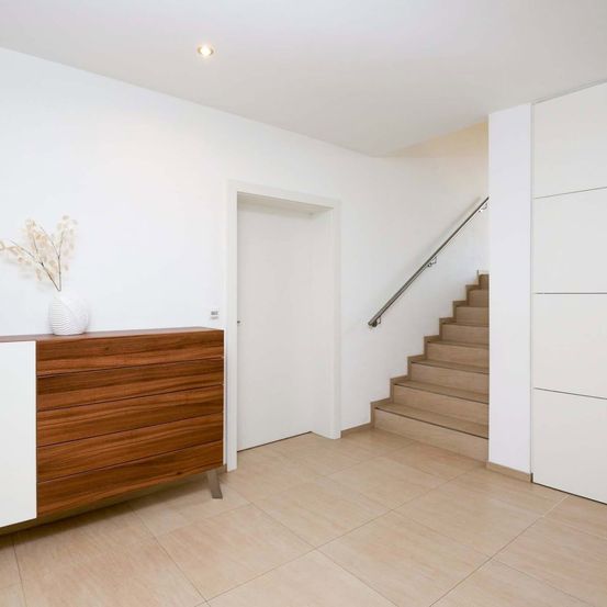 Ein modernes minimalistisches Interieur mit weißen Wänden, einem Holzschrank und einer Treppe, die in den oberen Stock führt. Ein weißer Vase mit getrockneten Blumen steht auf dem Schrank.