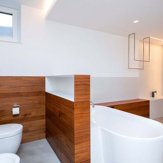 Modernes Badezimmer mit hölzerner Wand, weißer Badewanne, Waschbecken, Toilette und silbernen Armaturen. Ein Fenster und Deckenleuchten sorgen für Licht.